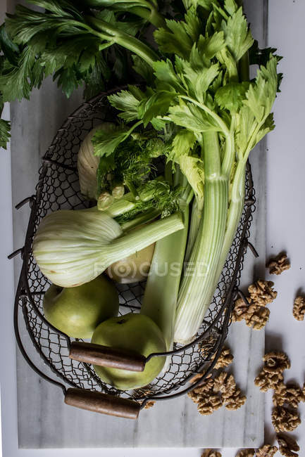 From above metal basket with apples, celery, fennel bulbs and walnuts arranged on board against white background — Stock Photo