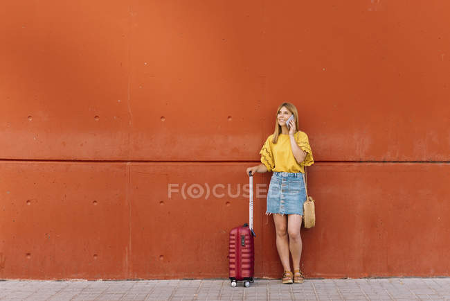 Молода жінка туристка з валізою розмовляє на мобільному телефоні, спираючись на червону стіну — стокове фото
