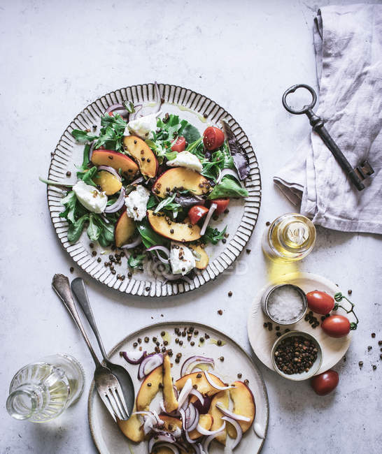 De cima pratos com saladas gourmet feitas de pêssegos, cebola vermelha, queijo, óleo e pimenta preta na mesa — Fotografia de Stock
