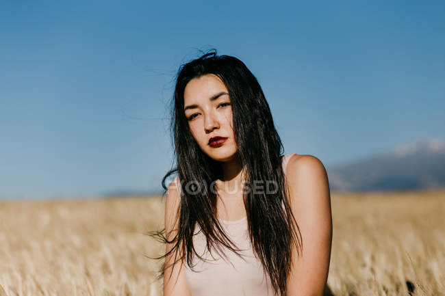Hermosa hembra asiática mirando a la cámara mientras está de pie sobre el fondo borroso del prado en el día ventoso en la naturaleza - foto de stock