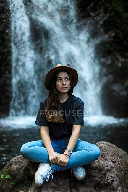 Fröhliche Reisende mit Hut lächelt und blickt in die Kamera, während sie auf nassen Felsbrocken in der Nähe des Wasserfalls sitzt — Stockfoto