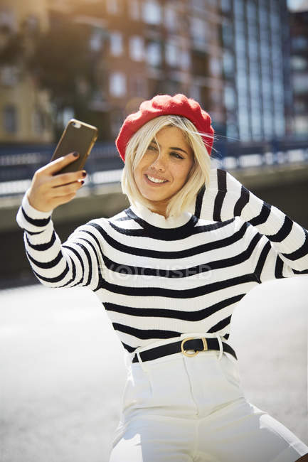 Jeune femme souriante en bonnet rouge français, chemisier rayé et short blanc prenant des photos sur fond urbain — Photo de stock
