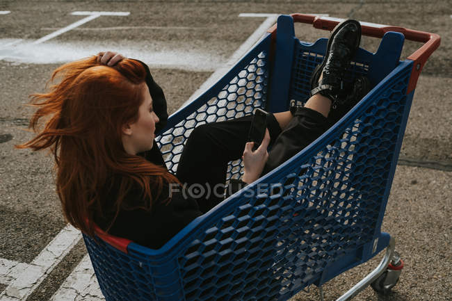 Улыбающаяся женщина, использующая смартфон в тележке для покупок на парковке — стоковое фото