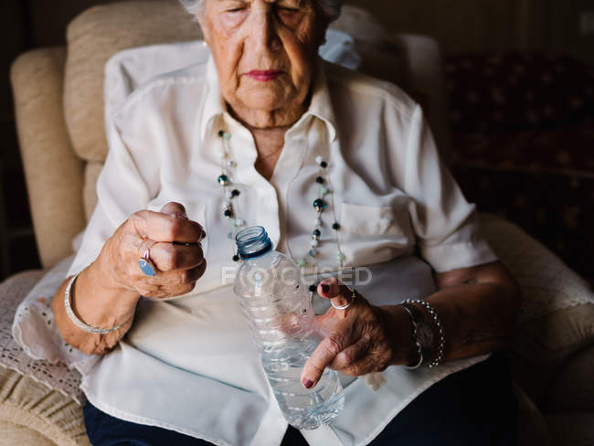 Seniorin im weißen Hemd trinkt Tabletten mit Wasser aus Flasche, sitzt auf Sessel und schaut in Wohnung weg — Stockfoto
