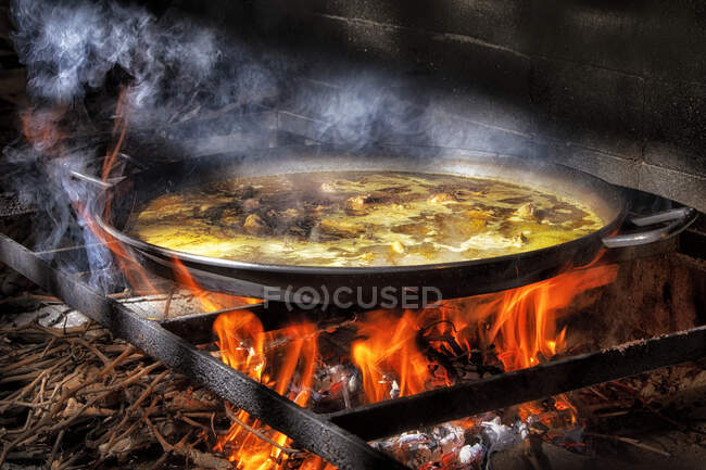 Grande casserole en fer avec bouillon bouillant pour cuisiner la paella sur un feu ouvert avec du bois — Photo de stock