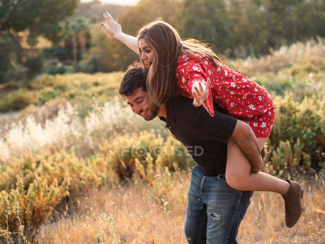 Cheerful couple playing piggyback while standing among yellow grass of countryside in summer day — Stock Photo