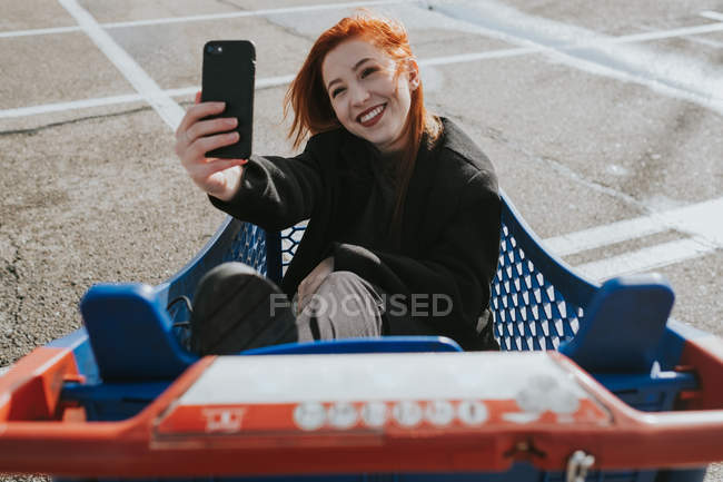 Приваблива молода жінка з рудим волоссям посміхається і приймає селфі сидить у синьому кошику покупок — стокове фото