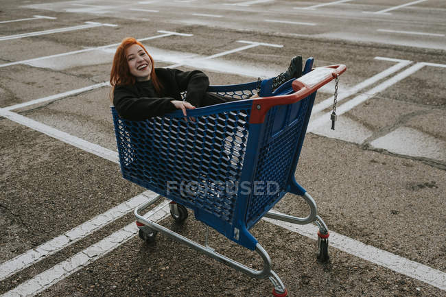Smiling woman sitting in shopping trolley in parking lot — Stock Photo
