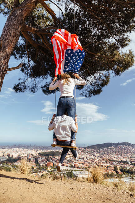 Dos chicas jóvenes con una bandera americana celebrando el 4 de julio en un columpio - foto de stock