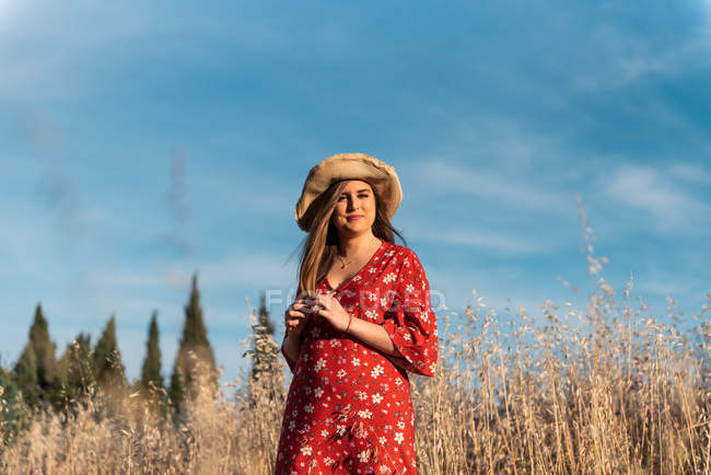 Приваблива усміхнена жінка в солом'яному капелюсі і червона сукня, що стоїть на дикому полі на тлі блакитного неба — стокове фото