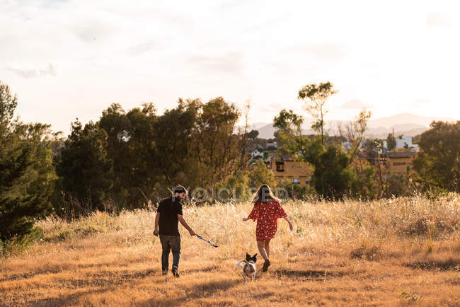 Casal alegre correndo com pequeno cão amigável entre grama amarela no parque — Fotografia de Stock