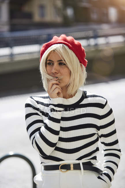 Jeune femme blonde en chemise rayée noire et blanche et casquette rouge française sur fond flou — Photo de stock