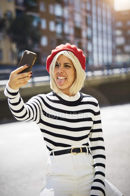 Молода усміхнена жінка у французькій червоній шапці, смугаста блузка і білі шорти роблять фотографії на міському фоні. — стокове фото