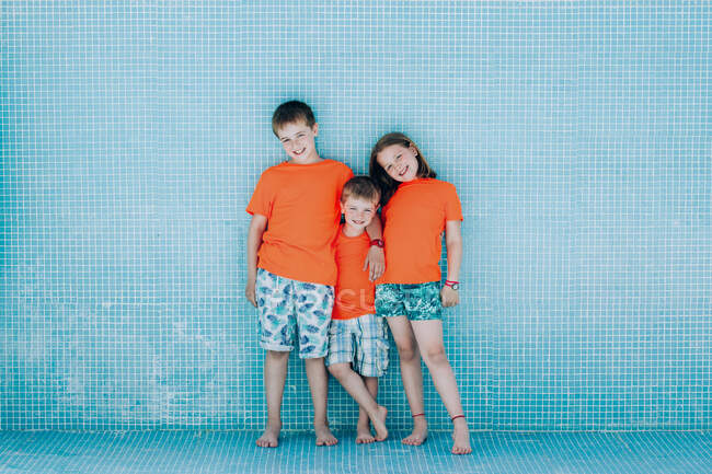 Niños en camisetas de color naranja brillante sonriendo y posando sobre el fondo de la pared de la piscina vacía - foto de stock