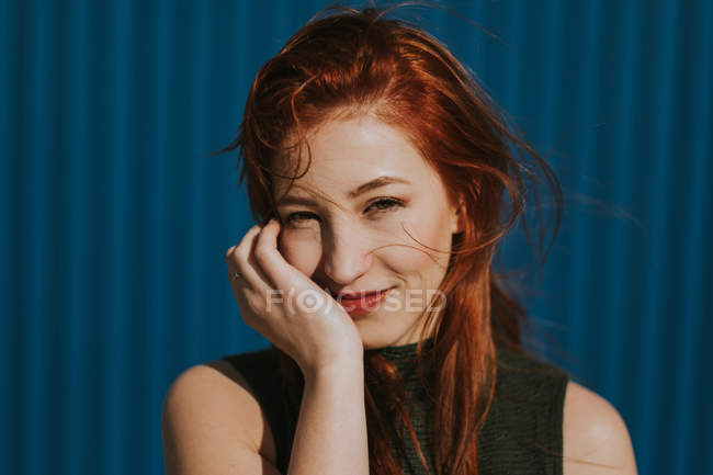 Приваблива молода жінка з рудим волоссям дивиться в камеру як зворушливе обличчя в сонячний яскравий день на блакитну стіну — стокове фото