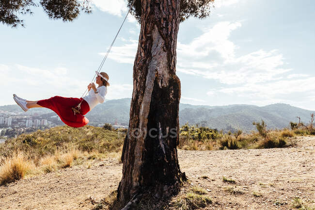 Chica divirtiéndose con falda roja y sombrero balanceándose mientras contempla la ciudad en el fondo - foto de stock