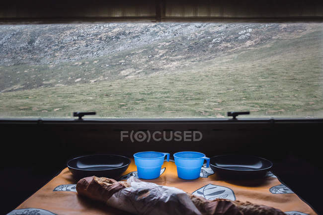 Pan de pan fresco puesto sobre la mesa cerca de tazas vacías y tazones contra ventana con vistas al terreno montañoso. - foto de stock