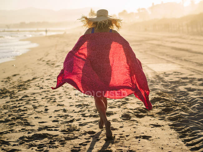 Невпізнанний жінка в капелюсі розмахуючи червоним шарфом і біжить вздовж узбережжя в Сонячний світлий день — стокове фото