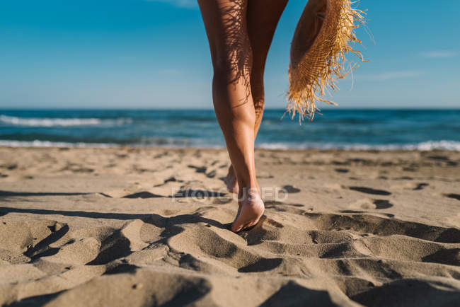 Visão traseira das pernas cortadas femininas com chapéu de palha na costa em luz solar brilhante — Fotografia de Stock