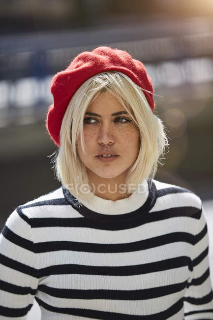 Mujer rubia joven con camisa blanca y negra a rayas y gorra francesa roja sobre fondo borroso - foto de stock