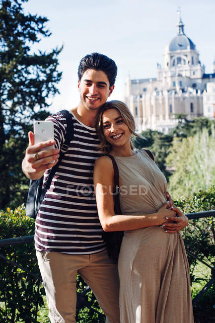 Joven hombre guapo tomando selfie con novia en hermoso jardín en el fondo del edificio histórico - foto de stock