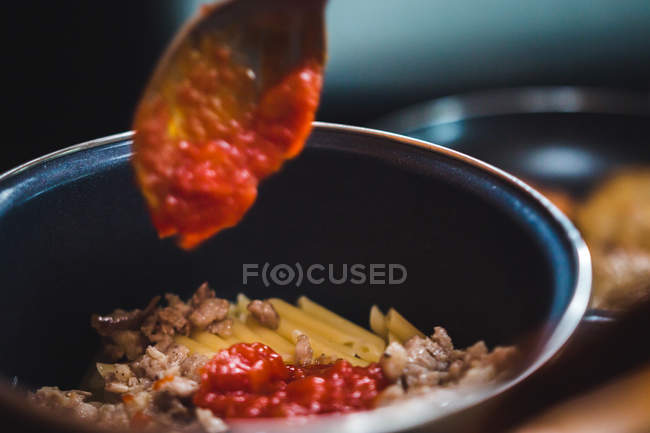 Cierre del cultivo inreconocible de pasta de cocinar a mano en una sartén con carne y salsa de tomate en la cocina. - foto de stock