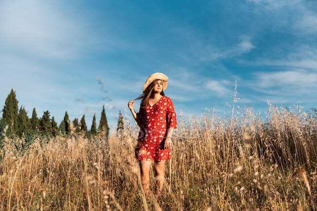 Sonriente mujer en sombrero de paja y vestido rojo de pie en el campo salvaje en el fondo del cielo azul - foto de stock