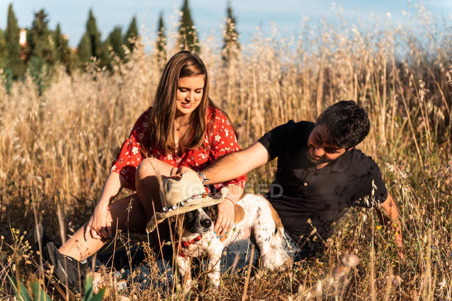 Cheerful couple sitting among high grass and stroking little dog in countryside — Stock Photo