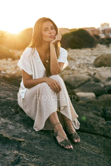 Элегантная молодая женщина сидит и улыбается на скале на закате — стоковое фото