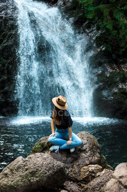 Cheerful female traveler in hat smiling and looking at camera while sitting on wet boulder near waterfall — Stock Photo