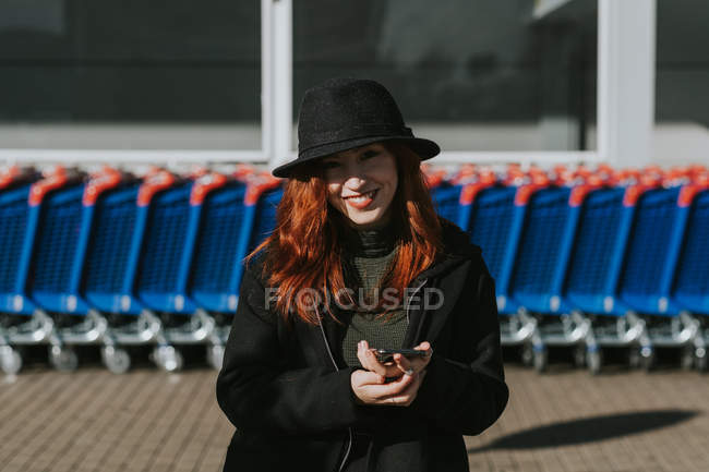 Jolie jeune femme aux cheveux roux en chapeau noir et veste souriant avec téléphone portable et regardant à la caméra dans le parking — Photo de stock