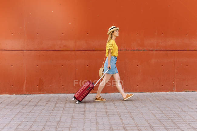 Молодая туристка идет с чемоданом против красной стены на улице — стоковое фото
