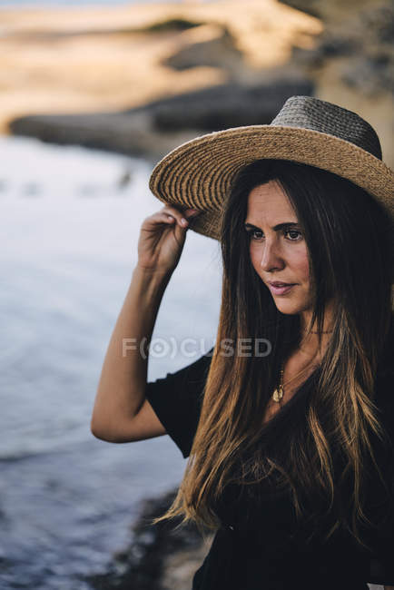 Jovem de cabelos longos mulher olhando para longe e segurando chapéu na praia — Fotografia de Stock