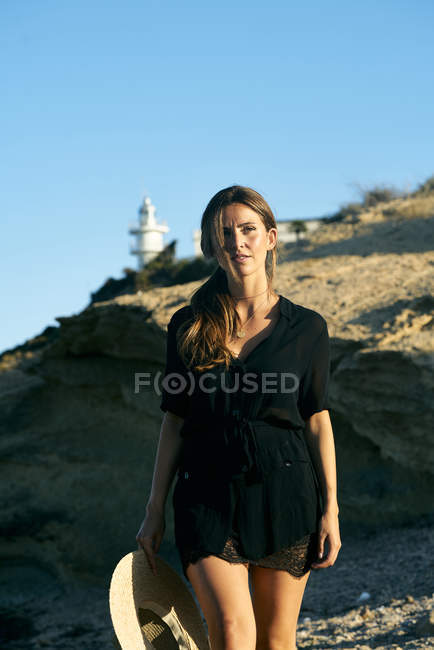 Giovane donna premurosa godendo il sole e guardando lontano sulla spiaggia con faro sullo sfondo — Foto stock