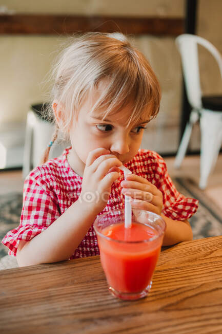 Appetitlich duftendes Glas roten Smoothie in den Händen eines entzückenden Kindes, das es am Tisch trinkt — Stockfoto