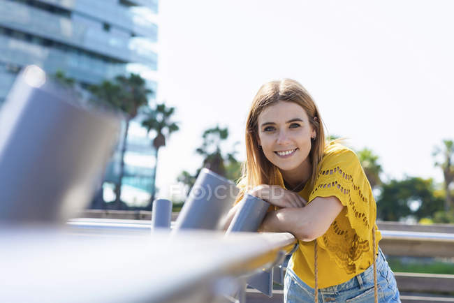 Porträt einer jungen lächelnden blonden Frau, die in die Kamera blickt, die am Geländer lehnt — Stockfoto