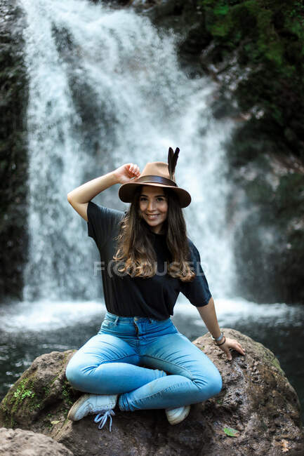 Весела жінка-мандрівник в капелюсі посміхається і дивиться на камеру, сидячи на мокрій валуні біля водоспаду — стокове фото