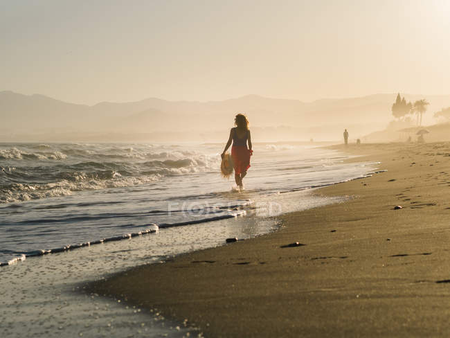 Visão traseira da mulher magra com chapéu e pareo andando ao longo da costa em luz solar brilhante — Fotografia de Stock