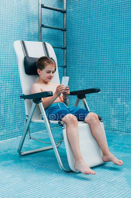 Male kid using smartphone while resting on chair in empty pool decorated by blue tiles — Stock Photo