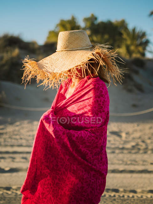 Mujer irreconocible en sombrero envuelto en una bufanda rosa en la orilla del mar en un día soleado y brillante - foto de stock
