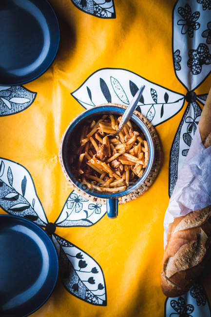 From above mug with tasty pasta placed on ornamental tablecloth near loaf of bread and empty bowls — Stock Photo