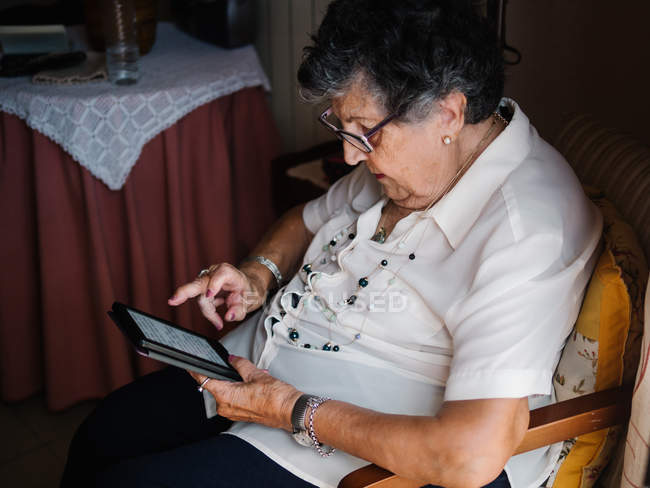 Mulher sênior em camisa e óculos sentados na poltrona e ler e-book no apartamento — Fotografia de Stock