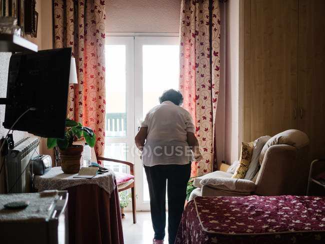 Rückansicht einer älteren Frau, die im Schlafzimmer zum Fenster geht — Stockfoto
