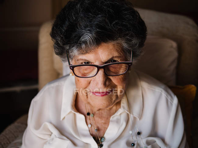 Retrato de mulher idosa feliz em camisa branca e com contas no pescoço olhando para a câmera em casa — Fotografia de Stock