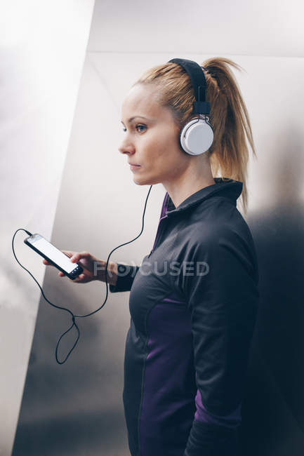 Joven rubia caucásica con ropa deportiva escuchando música con auriculares conectados a su smartphone - foto de stock
