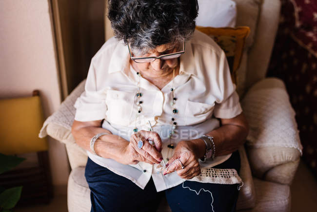 Elderly woman in blouse knitting crochet while sitting on armchair — Stock Photo