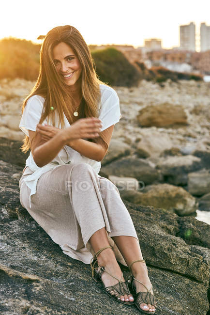 Елегантна молода жінка сидить і посміхається на скелі в природі на заході сонця — стокове фото