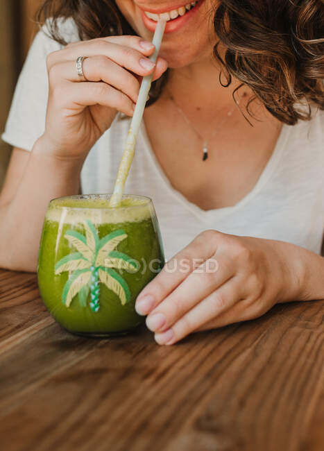 El vaso apetitoso perfumado del batido verde en las manos de la mujer bebiéndolo en la mesa - foto de stock