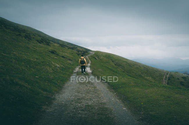 Вид сзади неузнаваемого самца с рюкзаком, стоящего на неровной дорожке на склоне холма на фоне пасмурного неба в природе — стоковое фото