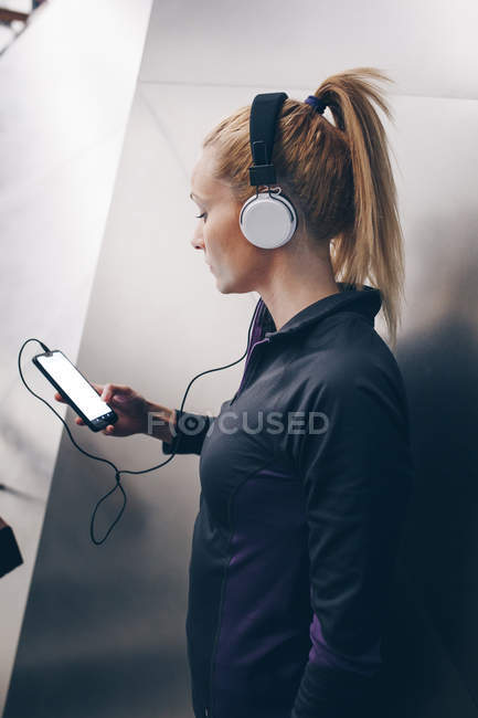 Junge blonde kaukasische Frau mit Sportbekleidung, die Musik mit Kopfhörern hört, die mit ihrem Smartphone verbunden sind — Stockfoto